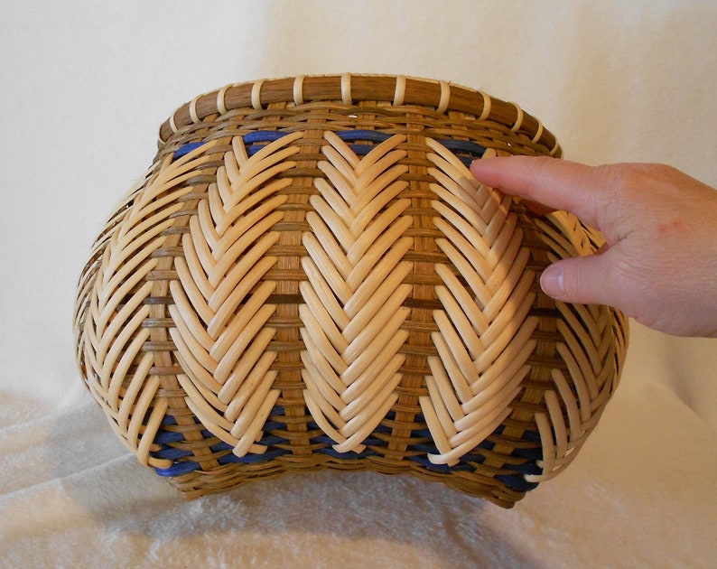 Puede incluir: Una cesta tejida con un patr&oacute;n de espiga en colores naturales y azules. La cesta es redonda y tiene una abertura ancha.