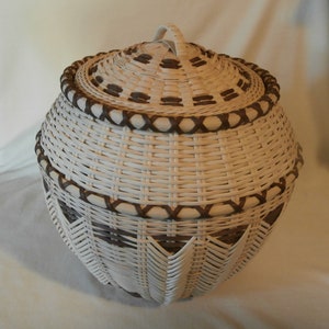 May include: A white wicker basket with a brown woven lid and handle. The basket has a decorative pattern of brown woven strips around the top and bottom.