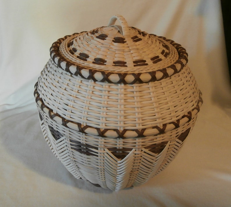 May include: A white wicker basket with a brown woven lid and handle. The basket has a decorative pattern of brown woven strips around the top and bottom.