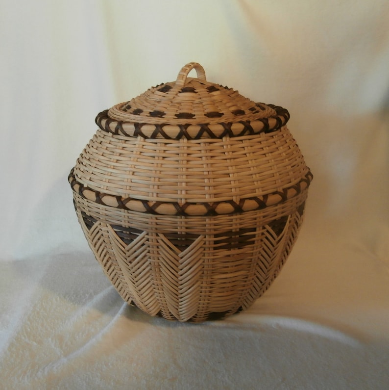 May include: A woven basket with a lid, featuring a brown and tan colour scheme. The basket has a decorative pattern of woven triangles on the bottom half.