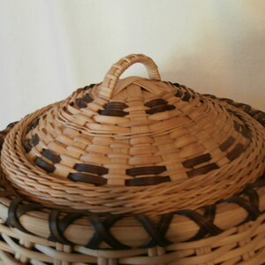 May include: A woven basket with a lid and a handle. The basket is made of natural materials and has a brown and tan colour scheme.