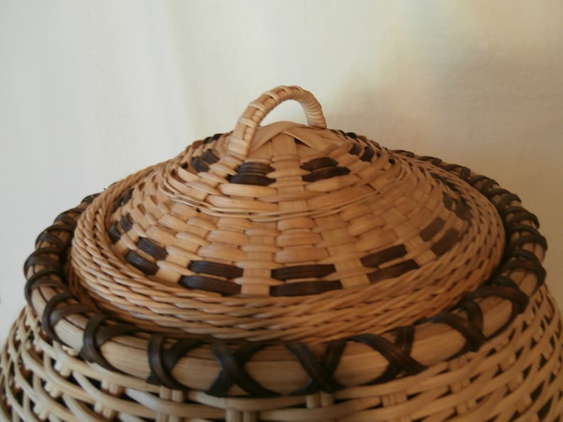 May include: A woven basket with a lid and a handle. The basket is made of natural materials and has a brown and tan colour scheme.