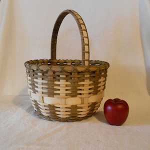 May include: A woven basket with a brown and white pattern and a wooden handle. The basket is sitting on a white surface next to a red apple.