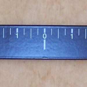 Op de afbeelding: Een zwarte spaakgewichtmeter met witte markeringen van 0 tot 8. De meter is gemarkeerd met "Spoke Weight" en heeft de tekst "Augusta Wheel Technology, 2002 Airport Road, Pageland, SC 29728, 843-672-5188" aan de zijkant gedrukt.