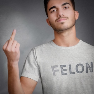 May include: A man wearing a grey t-shirt with the word "FELON" printed on it. He is giving the middle finger.