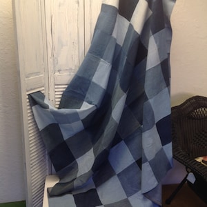 Puede incluir: Una colcha patchwork de mezclilla con un diseño de tablero de ajedrez en tonos azules, drapeada sobre una pantalla blanca con persianas. La colcha es un elemento de decoración del hogar. Dimensiones: aproximadamente 150 cm x 200 cm.