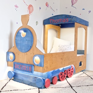 May include: A wooden train bed frame painted blue and brown with red wheels. The bed has a canopy with the name "Thomas" written on it. The bed has working lights on the front and a license plate that says "BK25818".