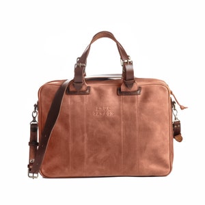 May include: A brown leather briefcase with a top handle and a shoulder strap. The bag has a zipper closure and a small pocket on the front. The bag has the text "KRUK GARAGE" embossed on the front.