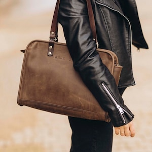 May include: A brown leather tote bag with a top handle and a zipper closure. The bag has the word "KRUK" embossed on the front.