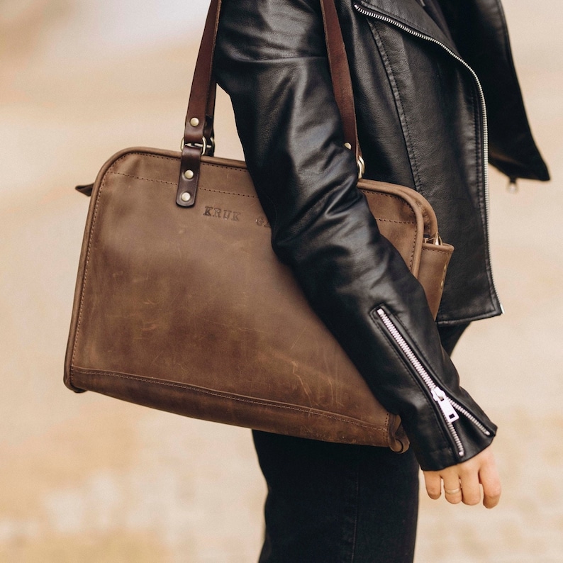 May include: A brown leather tote bag with a top handle and a zipper closure. The bag has the word "KRUK" embossed on the front.