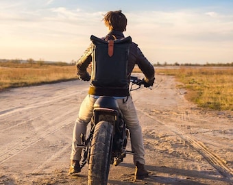 Puede incluir: Una persona conduciendo una motocicleta negra por un camino de tierra. La persona lleva una chaqueta de cuero marrón, una mochila negra y pantalones beige. La motocicleta tiene un asiento de cuero negro y un tubo de escape negro.