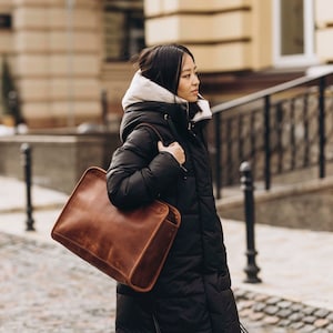 May include: A woman in a black puffer jacket carries a brown leather tote bag with a strap over her shoulder.