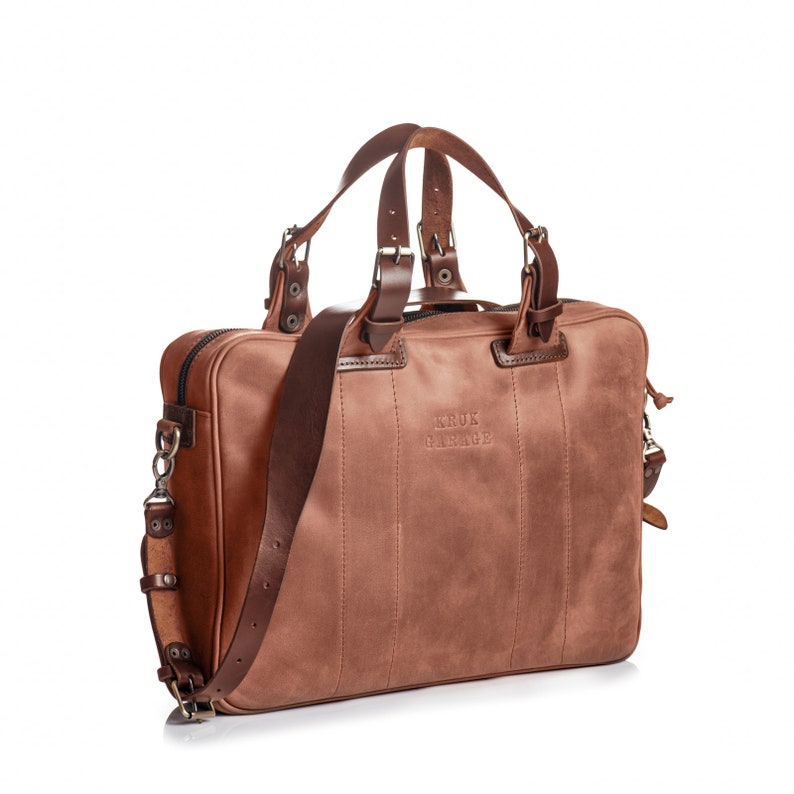 May include: A brown leather briefcase with a top handle and an adjustable shoulder strap. The bag has a zipper closure and a metal buckle. The bag has the text "KRUK GARAGE" embossed on the front.