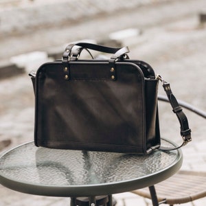 May include: A black leather tote bag with a top handle and a shoulder strap. The bag has gold-colored hardware and the text "THE CARRIER" embossed on the front.