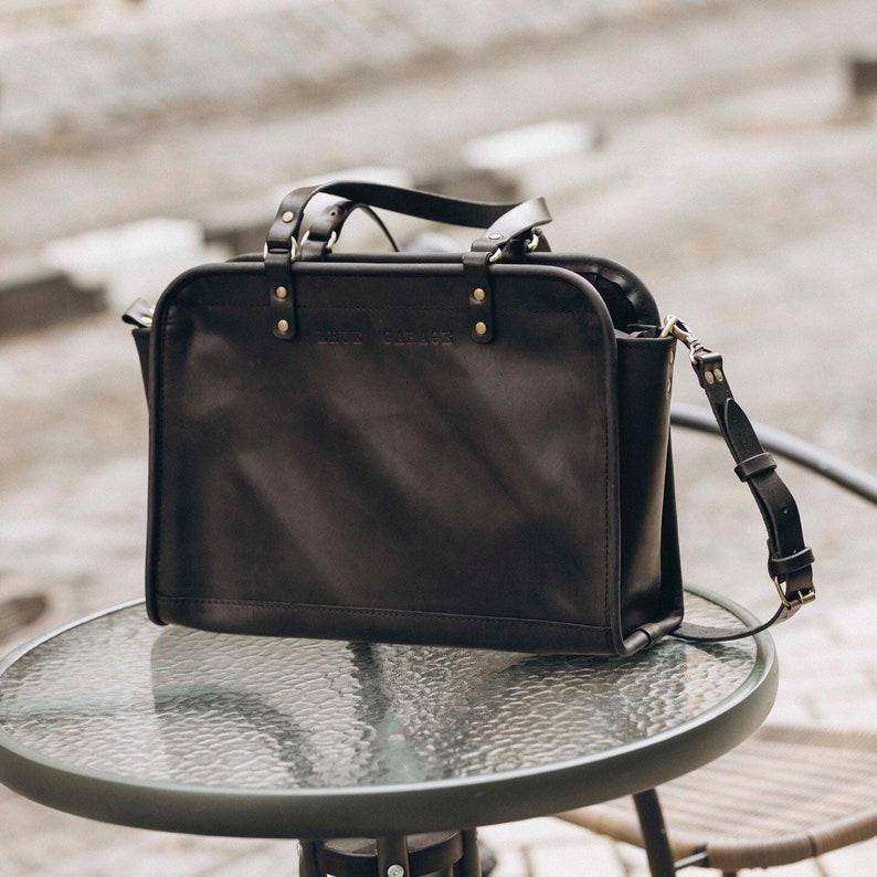 May include: A black leather tote bag with a top handle and a shoulder strap. The bag has gold-colored hardware and the text "THE CARRIER" embossed on the front.