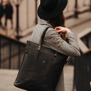 Könnte beinhalten: Eine schwarze Ledertasche mit Schulterriemen. Die Tasche hat eine rechteckige Form mit einer Vordertasche und wird von einer Person getragen, die einen grau gestreiften Blazer und einen schwarzen Hut trägt. Die Tasche hat goldfarbene Nieten.