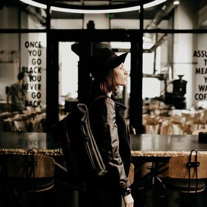May include: A woman wearing a black leather jacket and a black hat stands in a coffee shop. She is looking to the right. The text "ONCE YOU GO BLACK YOU NEVER GO BACK" is visible on the wall behind her.