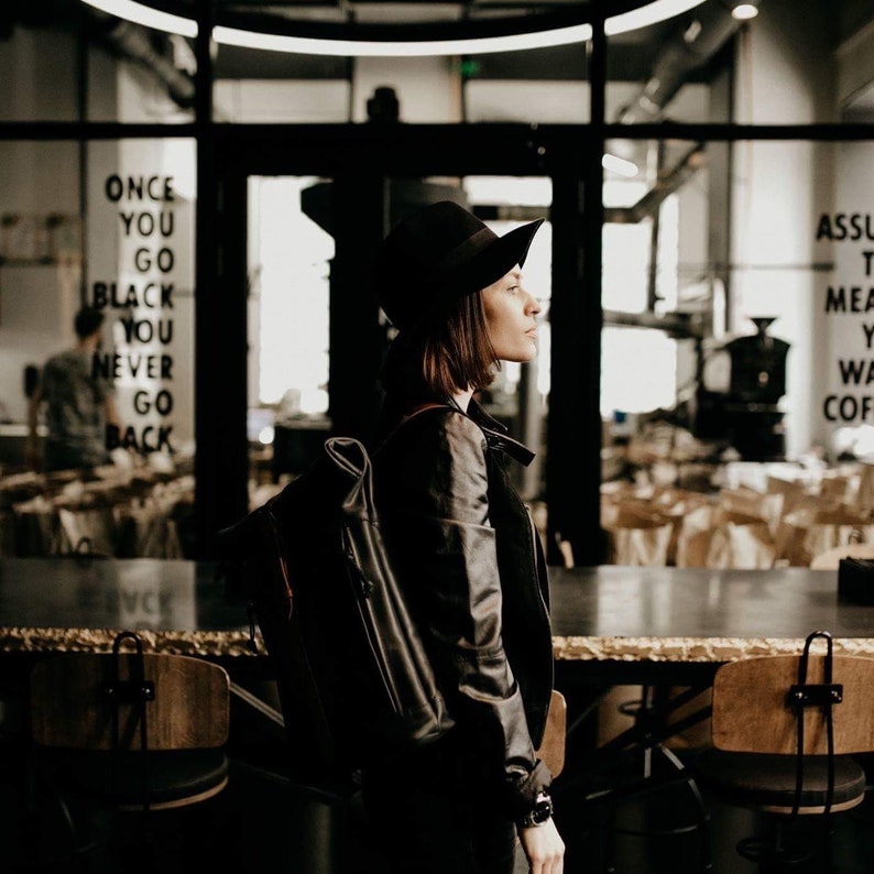 May include: A woman wearing a black leather jacket and a black hat stands in a coffee shop. She is looking to the right. The text "ONCE YOU GO BLACK YOU NEVER GO BACK" is visible on the wall behind her.