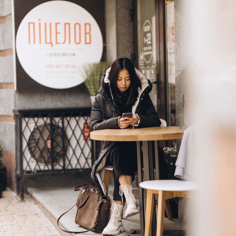 May include: A woman wearing a black and white winter coat sits at a table in front of a cafe. She is looking down at her phone. The cafe sign says "Пицелов" in Cyrillic script.