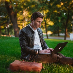 May include: A man wearing a black leather jacket and a white shirt sits on the grass, using a laptop computer. He has a brown leather bag next to him.
