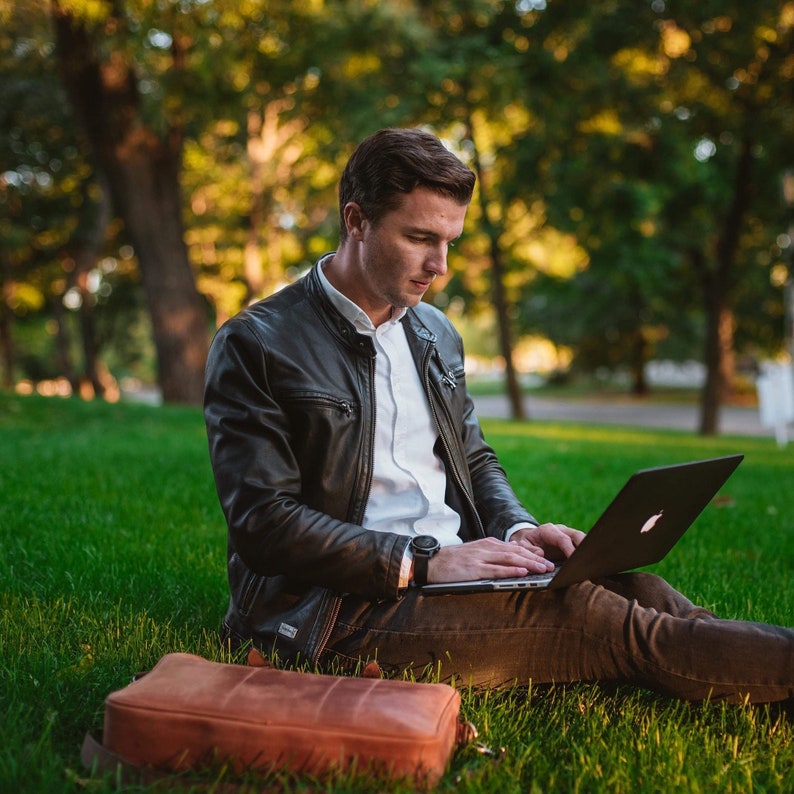 May include: A man wearing a black leather jacket and a white shirt sits on the grass, using a laptop computer. He has a brown leather bag next to him.