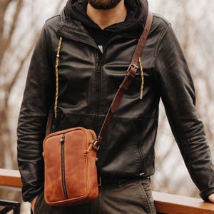 May include: A brown leather crossbody bag with a zip closure and a long adjustable strap. The bag is being worn by a person wearing a black leather jacket.