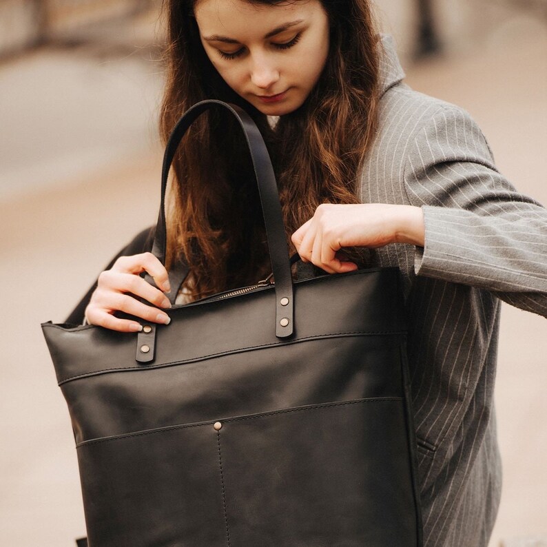 May include: A black leather tote bag with a zipper closure and gold hardware. The bag has a large main compartment and a smaller pocket on the front.