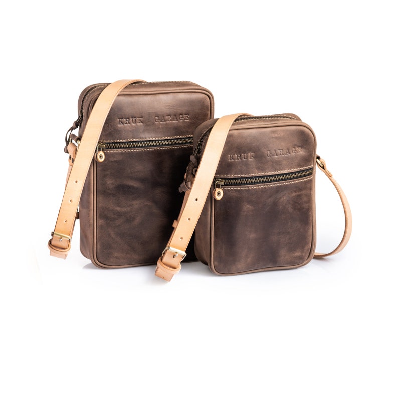 May include: Two brown leather crossbody bags with tan straps. The larger bag has the text "KRUK GARAGE" embossed on the front. The smaller bag has the same text embossed on the front.