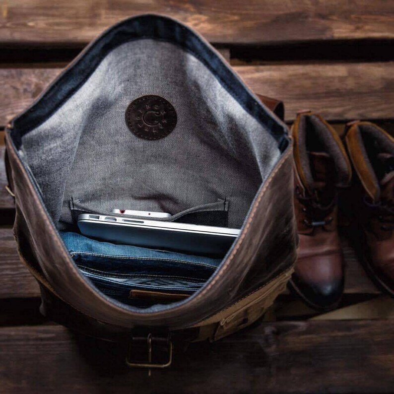 May include: A brown leather backpack with a flap closure and a zipped pocket. The backpack is open and a laptop computer is inside. The backpack is sitting on a wooden surface.