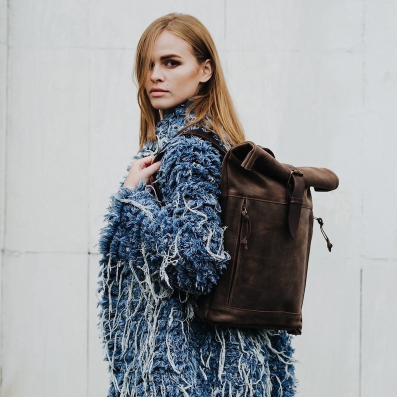 May include: A woman wearing a blue denim jacket with a brown leather backpack. The backpack has a roll-top closure and a buckle.
