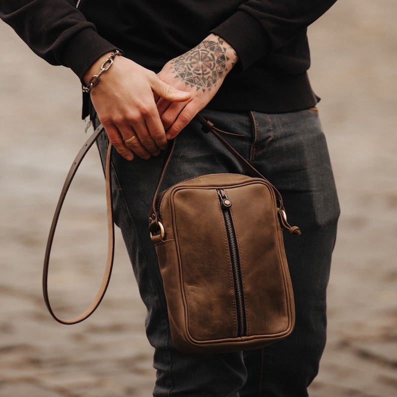 May include: A brown leather crossbody bag with a zip closure and a long strap. The bag is being worn by a person with a tattoo on their arm.