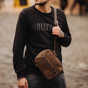 May include: A brown leather crossbody bag with a zip closure and a strap. The bag is being worn by a person wearing a black sweatshirt with the word "HOLLYWOOD" printed on the front.