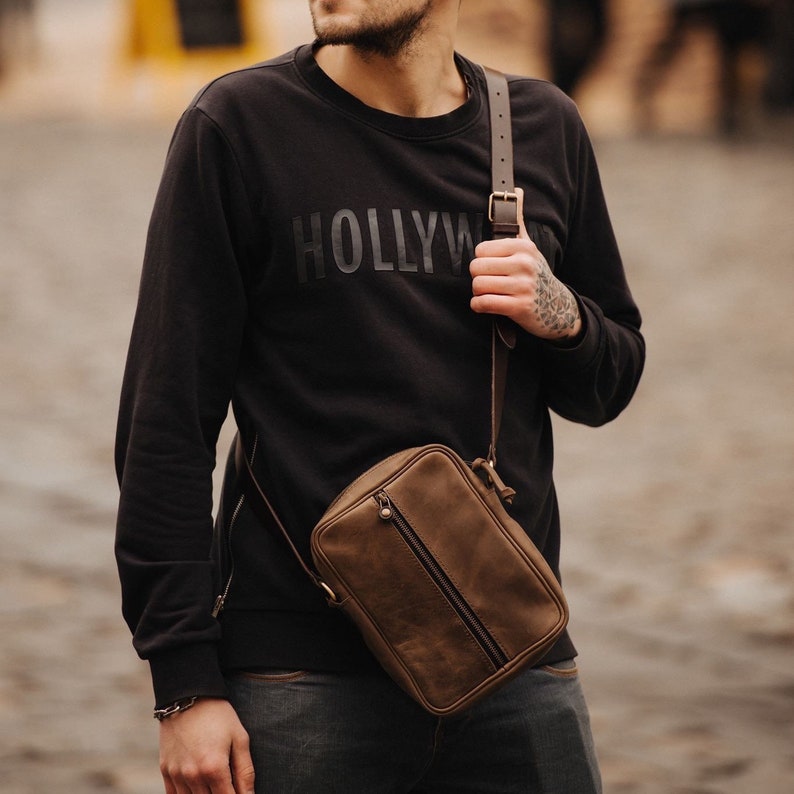 May include: A brown leather crossbody bag with a zip closure and a strap. The bag is being worn by a person wearing a black sweatshirt with the word "HOLLYWOOD" printed on the front.