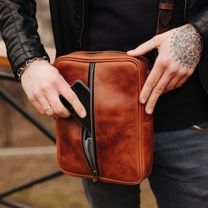 May include: A brown leather crossbody bag with a zipped pocket and a flap closure. A smartphone is visible inside the pocket.