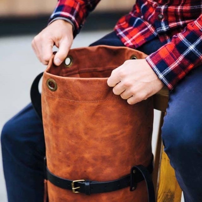 May include: A brown leather backpack with a black belt and buckle. The bag has brass eyelets around the top opening. The person holding the bag is wearing a red and black plaid shirt.