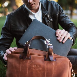 May include: A brown leather briefcase with a black laptop inside. The briefcase has a metal buckle and the text "KRUK GARAGE" embossed on the front.