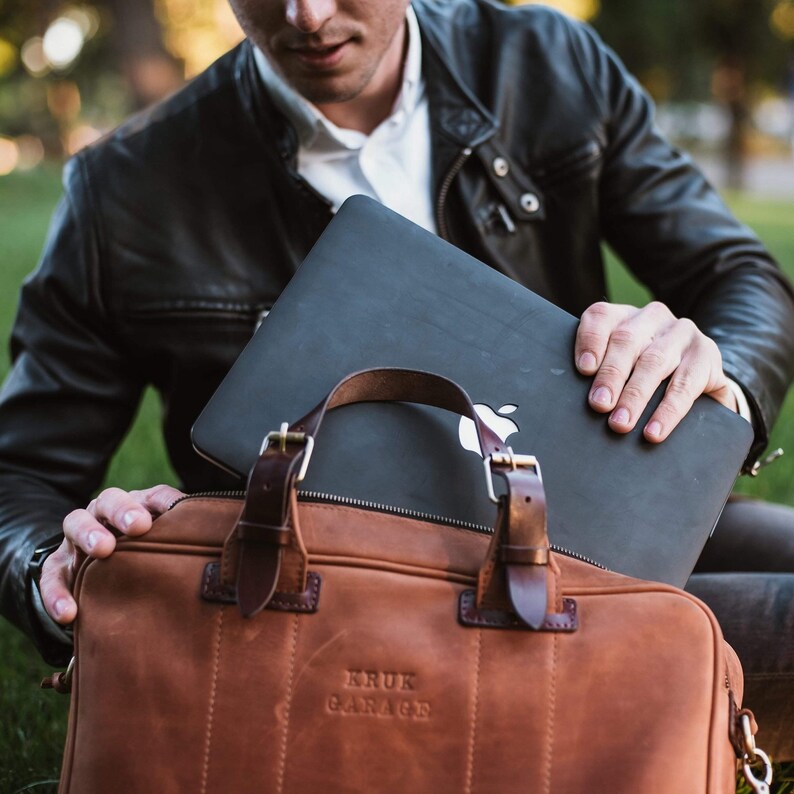 May include: A brown leather briefcase with a black laptop inside. The briefcase has a metal buckle and the text "KRUK GARAGE" embossed on the front.