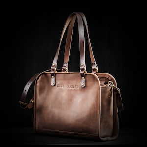 May include: A brown leather tote bag with two top handles and a shoulder strap. The bag has a zippered closure and a front pocket. The bag has the text "KRUK GARAGE" embossed on the front.