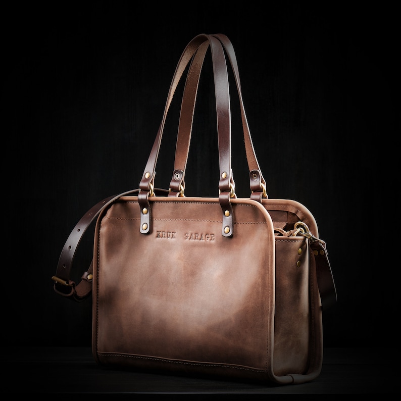 May include: A brown leather tote bag with two top handles and a shoulder strap. The bag has a zippered closure and a front pocket. The bag has the text "KRUK GARAGE" embossed on the front.