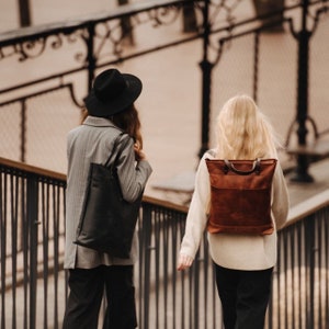 May include: Two people walking down a street, one wearing a black hat and carrying a black tote bag, the other wearing a white sweater and carrying a brown leather backpack.