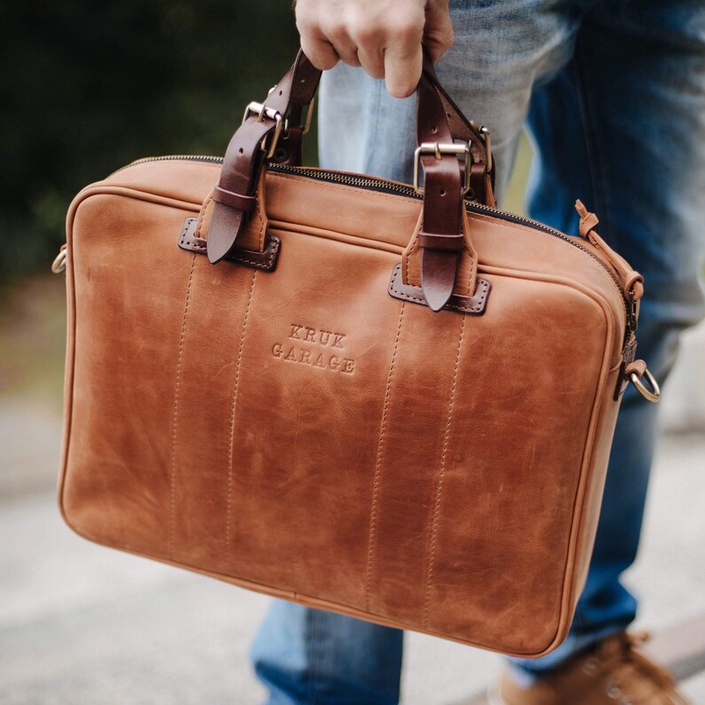 May include: A brown leather briefcase with a zipper closure and a top handle. The briefcase has a  "KRUK GARAGE" embossed logo on the front.