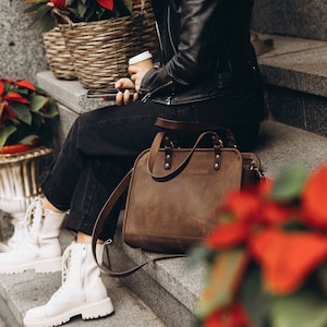 May include: A brown leather tote bag with a long strap, worn by a person wearing black pants and a black leather jacket. The bag has a top handle and a zipper closure.