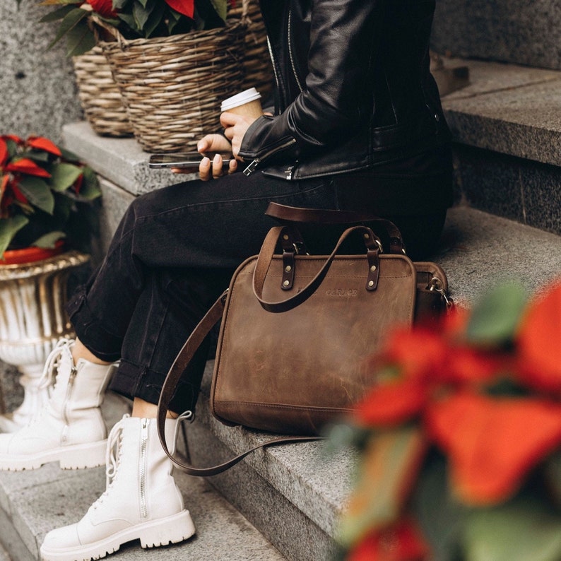 May include: A brown leather tote bag with a long strap, worn by a person wearing black pants and a black leather jacket. The bag has a top handle and a zipper closure.