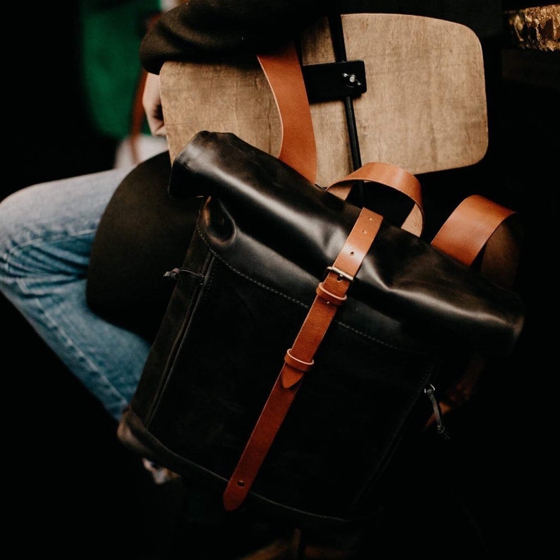 May include: A black leather backpack with brown leather straps and buckles. The backpack has a roll-top closure and is being worn by a person sitting on a wooden chair.