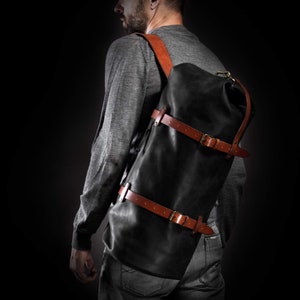 May include: A black leather backpack with brown leather straps and metal buckles. The backpack has a roll-top closure and a single shoulder strap. The bag is worn over the shoulder and across the back. The background is black.