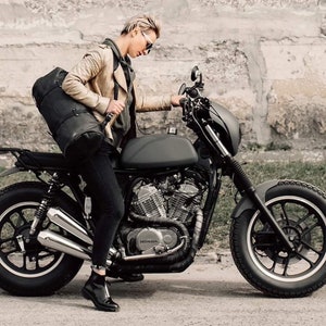 May include: A woman in a brown leather jacket and black pants sits on a black motorcycle. The motorcycle has a black leather saddlebag, a black seat, and a black engine. The motorcycle has a chrome exhaust pipe and a chrome Honda logo on the engine. The motorcycle has a black front fender and a black rear fender.