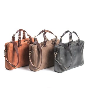May include: Three leather briefcases in different colors: brown, gray, and black. Each briefcase has a top handle and a shoulder strap. The briefcases are made of a textured leather and have a simple, classic design.