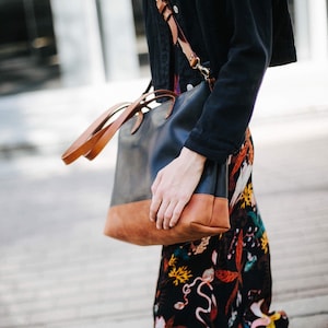 Może przedstawiać: Czarno-brązowa skórzana torba typu tote z długim brązowym paskiem skórzanym. Torba jest noszona na ramieniu osoby w czarnej kurtce i kwiecistej spódnicy.
