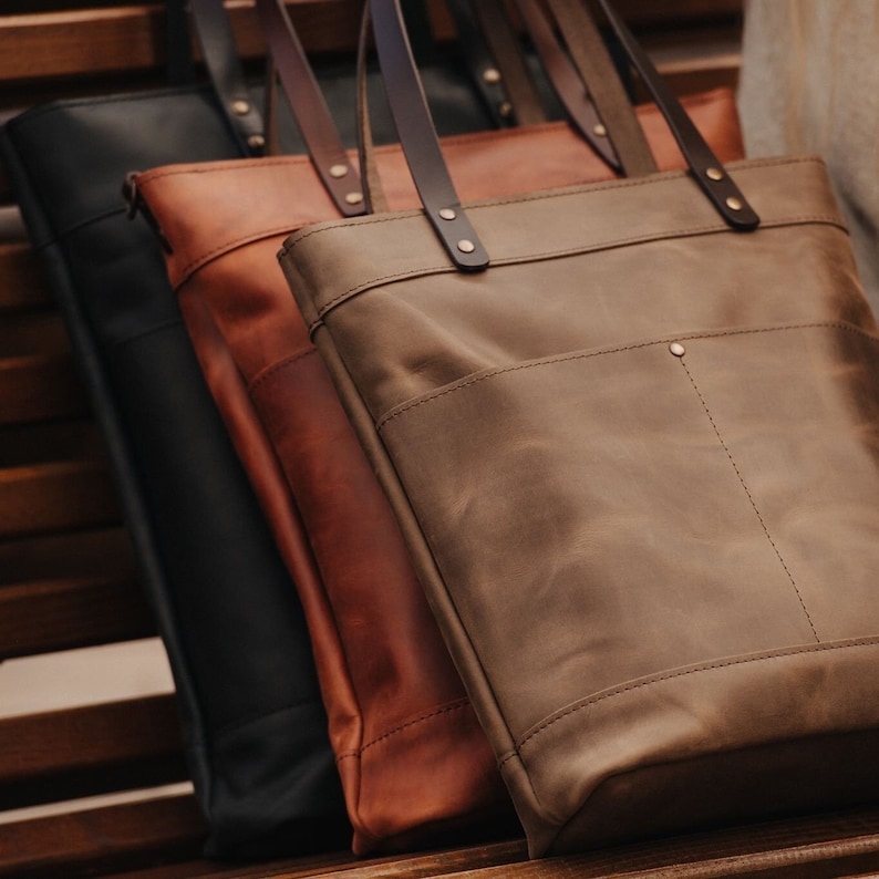 May include: Three leather tote bags in different shades of brown. The bags have a simple design with a single pocket on the front and a top handle.