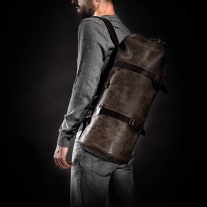 May include: A large, dark brown leather duffel bag with adjustable straps and buckles. The bag is worn over the shoulder and has a cylindrical shape. The leather has a distressed appearance, and the bag appears to be well-crafted.